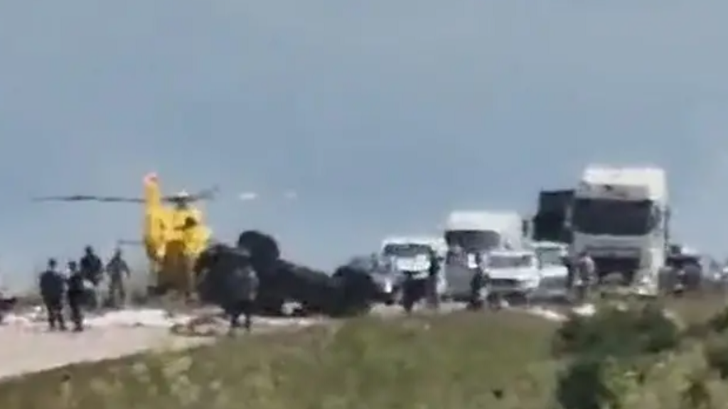 An image of the scene after the crash. It is blurry but a yellow air ambulance can be seen at the scene and a dark black vehicle can be seen on its roof. After vehicles and traffic are seen in the distance.
