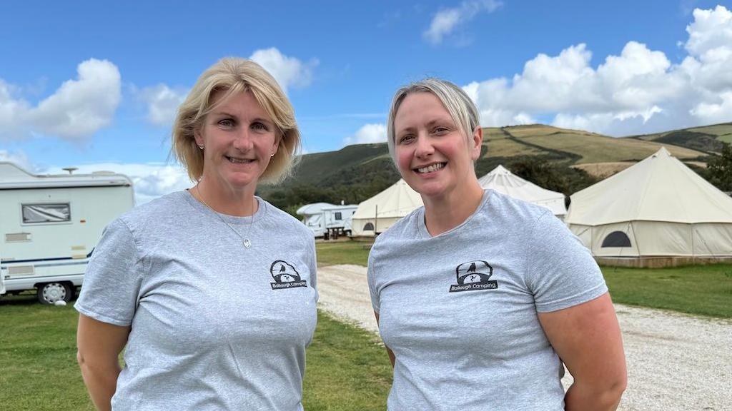 Two women are pictured wearing grey T-shirts. On the left is Cheryl Curphey, a woman with short blonde hair, and Zoe Huxham is pictured on the right. She also has blonde hair which is tied back. They are both smiling directly at the camera. There are tents and a hill in the background.
