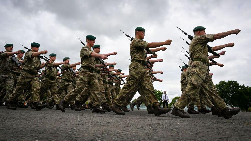 Royal Navy personnel are marching in unison holding a sharp spear on an overcast day