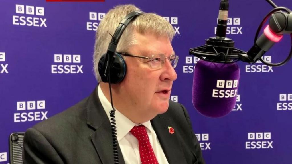 Roger Hirst, wearing headphones talking into a 91ȱ Essex microphone, with a 91ȱ Essex backdrop behind him