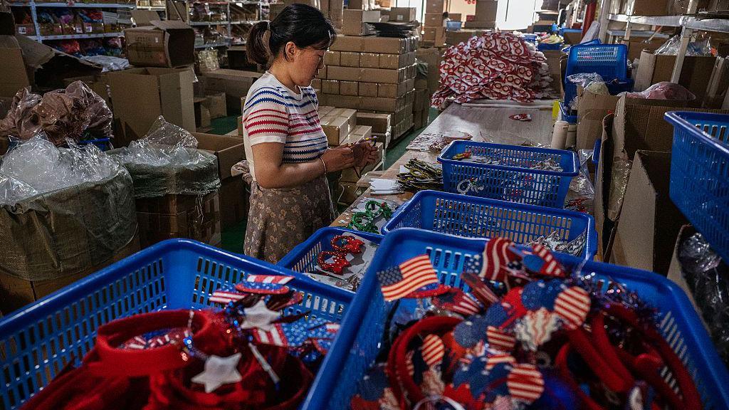 A worker makes festive goods for export at a factory on April 28, 2025 in Yiwu, Zhejiang Province, China. Pictured is a woman in a striped shirt and brown printed skirt standing in front of blue baskets filled with red American flags and other memoribilia.