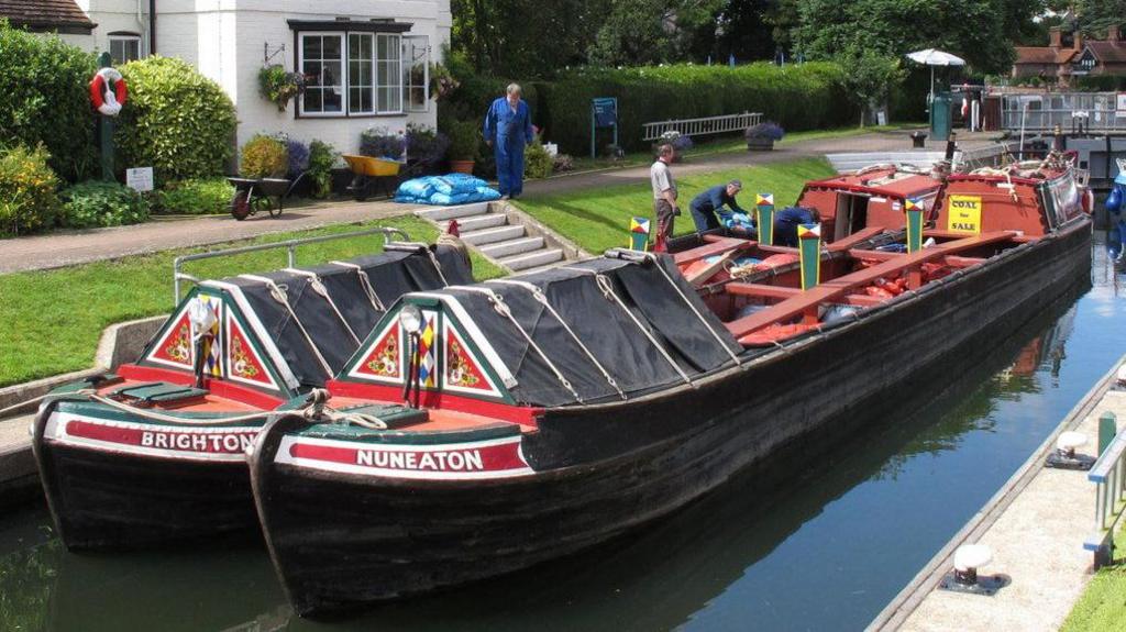 A narrowboat painted in red with NUNEATON in white lettering on the front is lying alongside a much shorter craft similarly painted with the word BRIGHTON on the side. The boats have covered canopies at the front. There are people standing on the bank which is mostly grass with some stone steps up to a white waterside cottage.