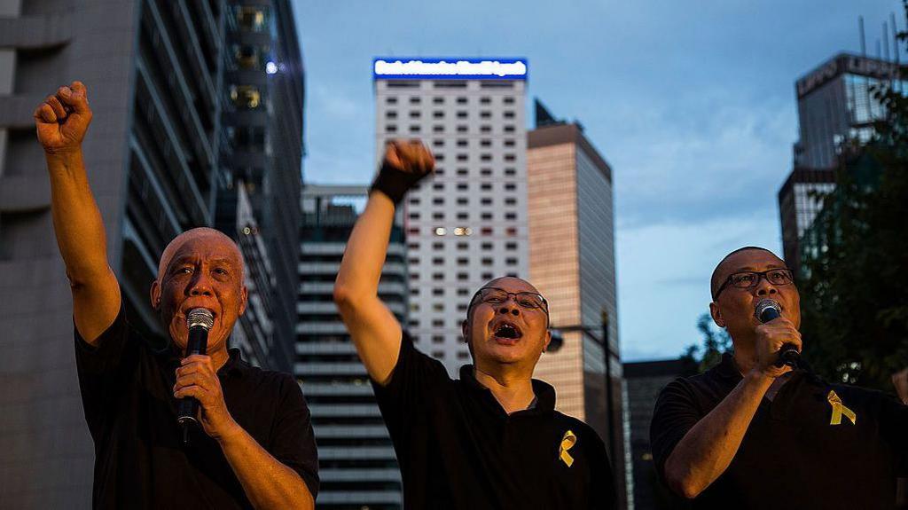 Democracy activists Chu You Ming (L), Benny Tai (C) and Chan Kin Man (R), speak on the stage after a march on September 14, 2014 in Hong Kong. They are wearing black t-shirts with golden ribbon pinned to them as they raise their fists in the air. 