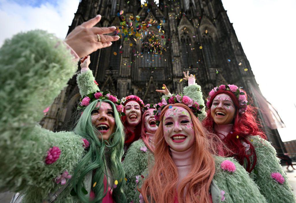 Women dressed in green outfits, pink and white face paint, and pink flowers in their hair, cheer in front of Cologne Cathedral while throwing confetti in the air