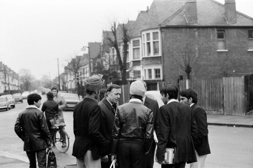 Two male detectives speak to a small group of people, including two men wearing turbans and two schoolboys in blazers, carrying books. The house where Eve Stratford died is in the background and another boy walks past and someone on a bicycle speaks to another police officer.