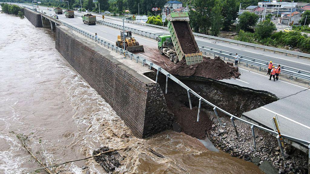 A bulldozer and truck beside a collapsed section of a road. Brown water is flowing in a canal beside the road.