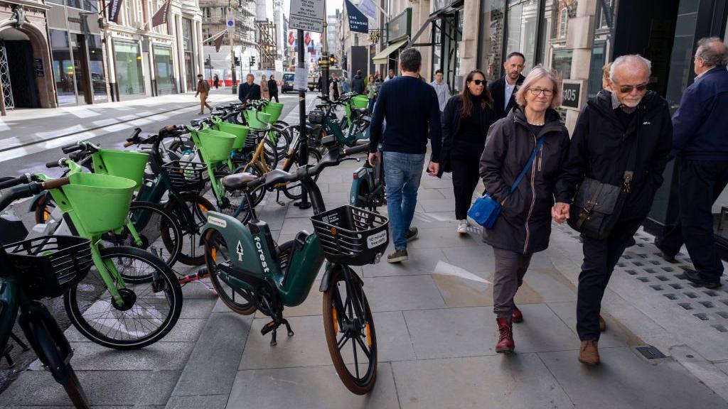 Lime and Human Forest e-bikes parked across the pavement along Bond Street, London. People walk past avoiding the bikes.