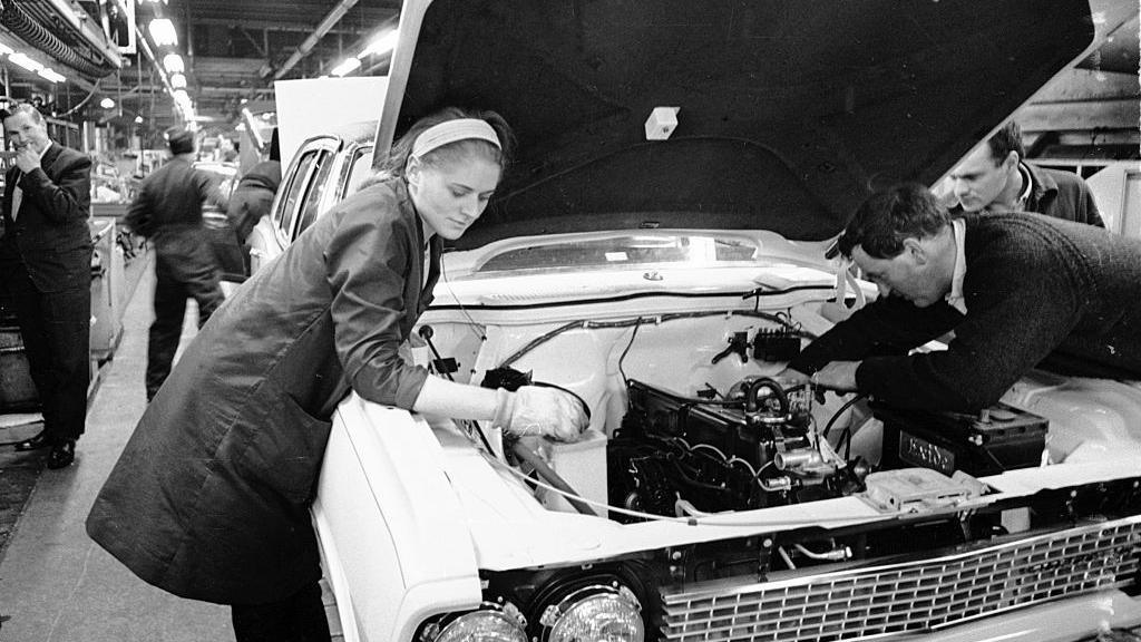 A black and white photo of workers helping to build a Vauxhall Victor. A woman wearing a headband and technician's coat is working on a car engine.