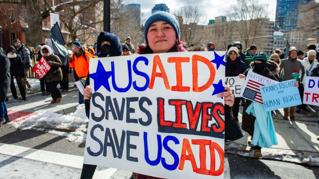 Woman holds sign that reads "USAID SAVES LIVES SAVE USAID"