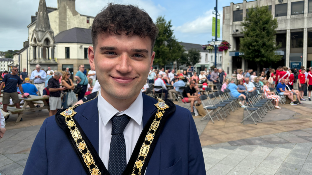 Man wearing mayoral chain and suit smiling into the camera. Crowd of people and rows of plastic chairs in a town square behind