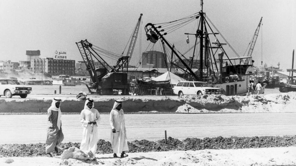 A black and white image of Dubai's port in 1977, with men in Arab dress in the foreground and behind them a few cranes