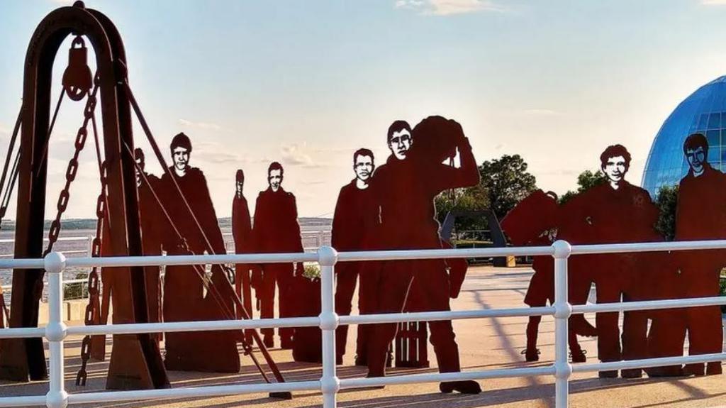 Steel sculpture depicting the crew of a trawler behind a white railing. Silhouette figures are in work poses from a fishing dock.