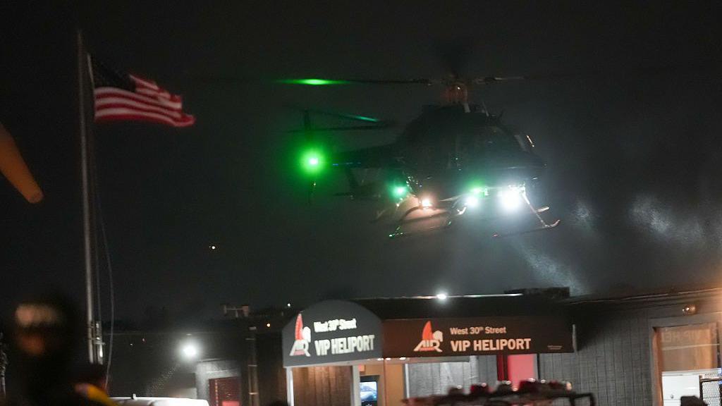 A helicopter flying in darkness with the US flag on a pole to the left and two "VIP Heliport" signs