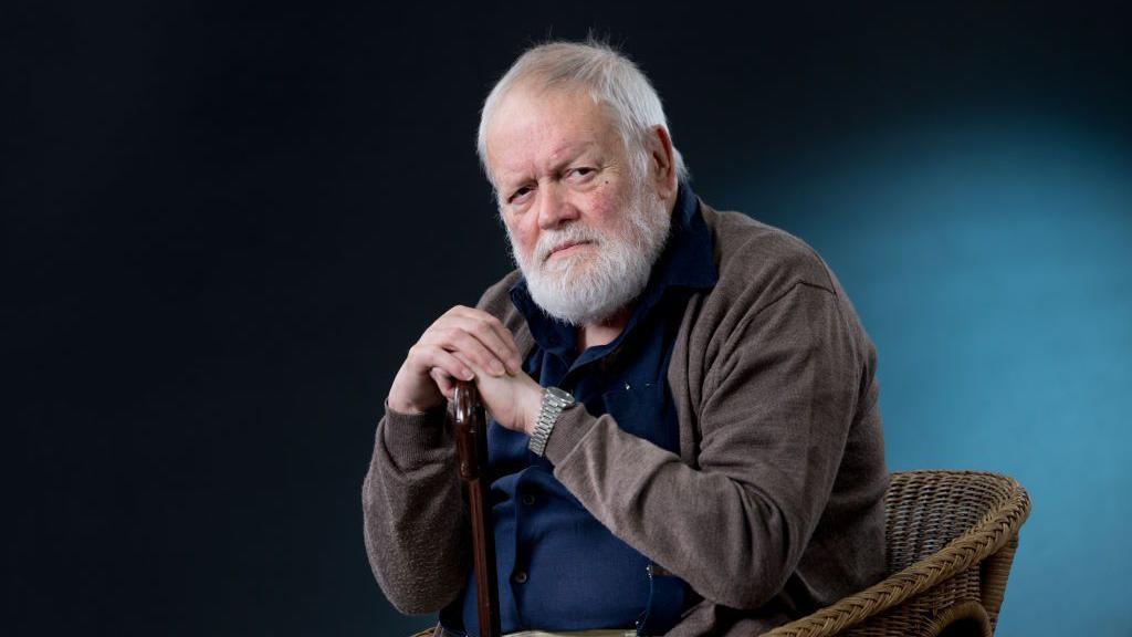 Longley, wearing a brown cardigan over a dark blue shirt, sits in a wicker chair resting his hands on a brown wooden walking stick.