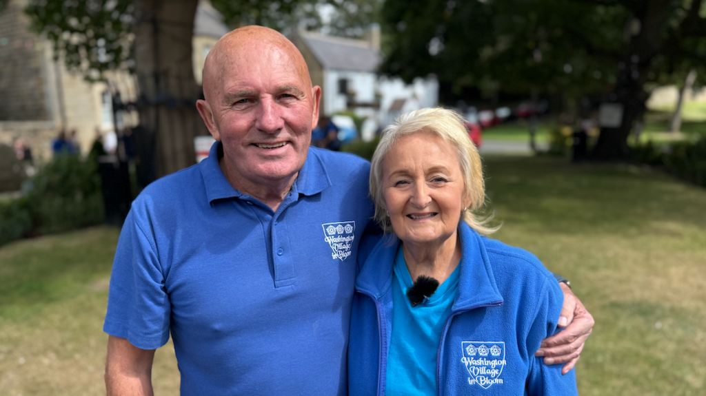 Joan Atkinson, right, with blonde and white hair and wearing a blue shirt with a blue fleece, which features a Washington Village in Bloom logo, stands next to partner Ron, who has no hair and is wearing a blue polo shirt with the same logo.