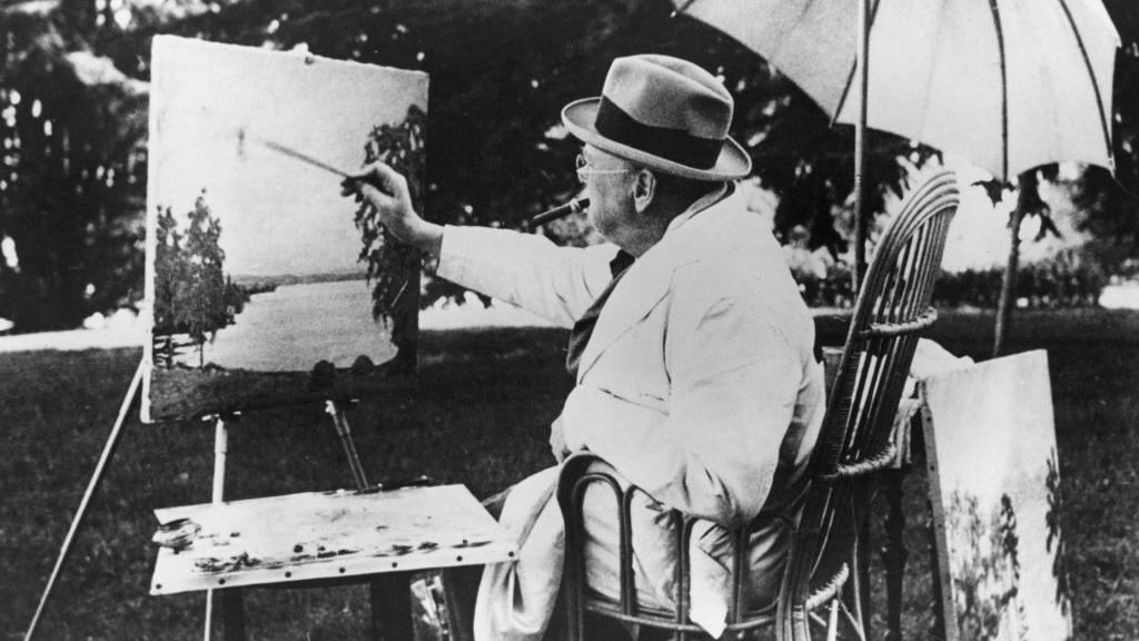 A black and white photo of Winston Churchill sitting on a chair, under an umbrella and painting.