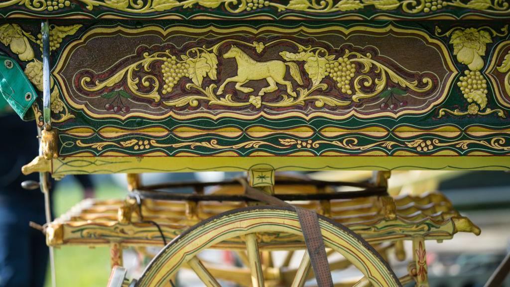 A close-up of the wheel and carriage of a traditional travellers' caravan, a horse, grape and leaf motif painted in gold. The caravan appears to be parked in a field, and a man's leg is blurred next to it.