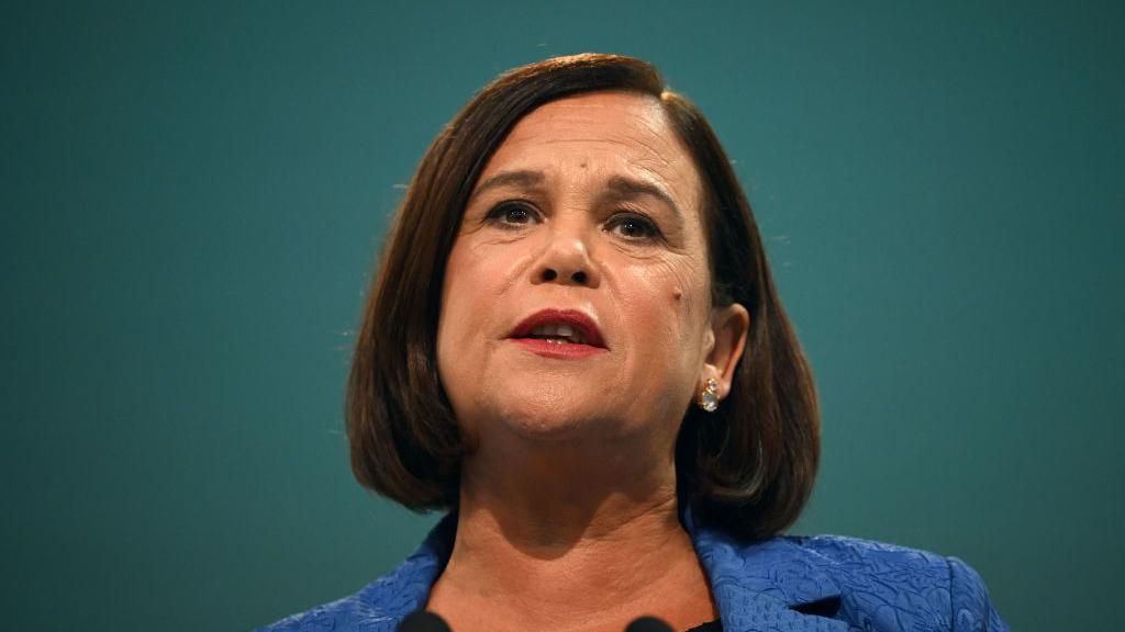 Mary Lou McDonald - a woman with shorter, brown hair stands against a green background as she delivers a speech. She is wearing a blue, patterned suit jacket.