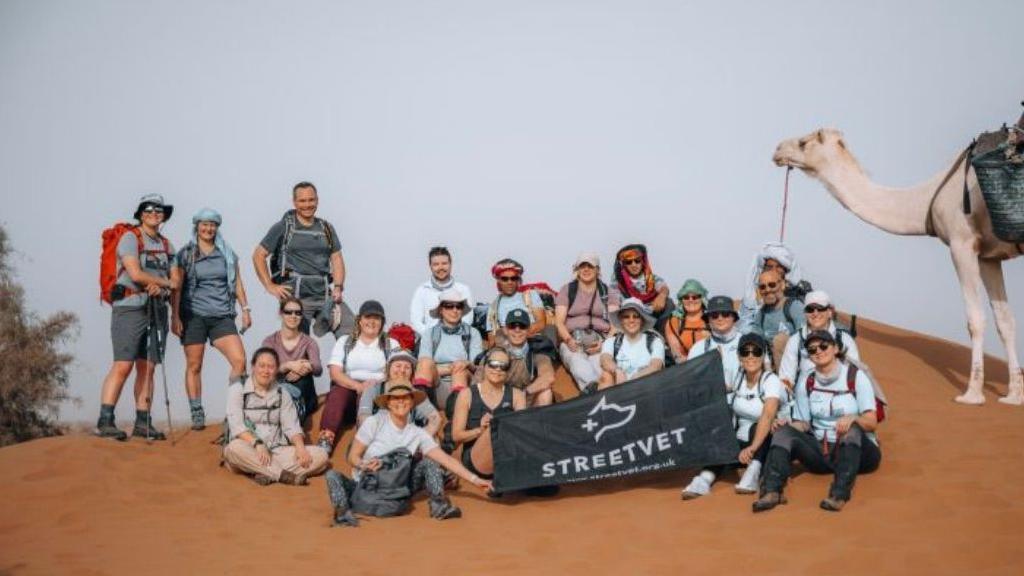 A group of men and women, some seated on the floor, some standing in the desert holding a black sign which reads StreetVet and there is a camel on the right-hand side