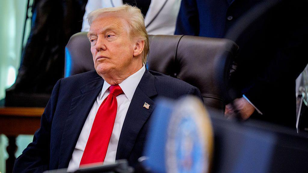 Donald Trump sitting at a desk, wearing a suit and red tie
