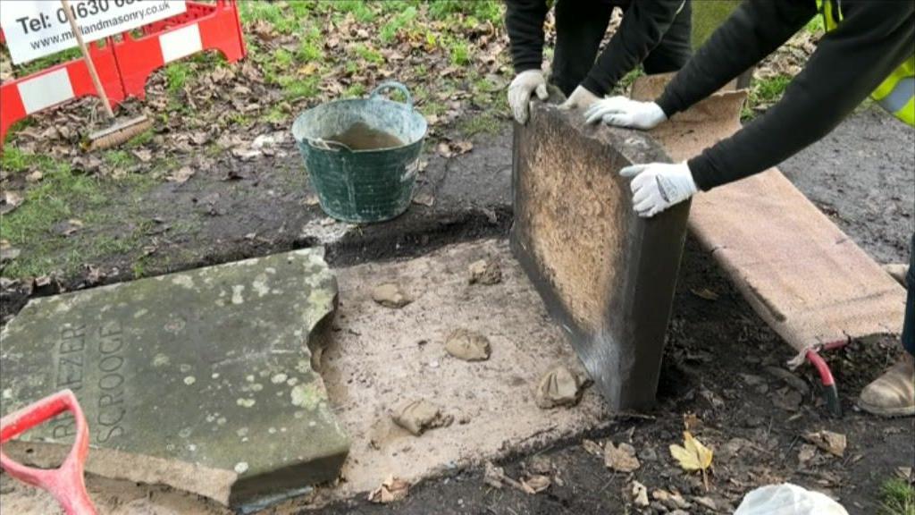 Two people standing up a large piece of the gravestone. Lying in front of it is another large piece of the stone, which says "Emenezer Scrooge". There are buckets and equipment around the stone.