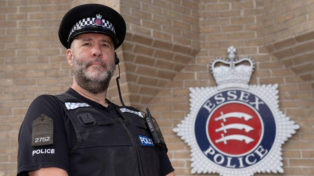 PC Dave Bishop is pictured outside the Colchester Police Station. He is standing in his police uniform outside the red brick building. The Essex Police logo is on the wall behind him.