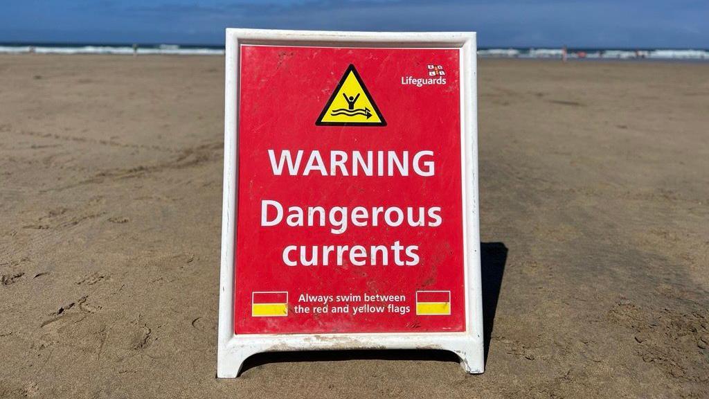 A red RNLI warning board on the sand at a beach. It has the words 'WARNING Dangerous currents' and below it 'Always swim between the red and yellow flags'. Either side of the text there are two red and yellow flags.