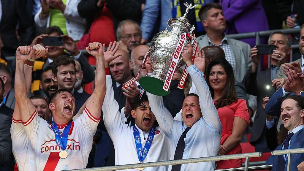 Hull KR coach Willie Peters, in a pale shirt and black tie, lifts the Challenge Cup above his head in front of the Royal Box at Wembley, with players alongside him in white and red shorts joining in the celebration.
