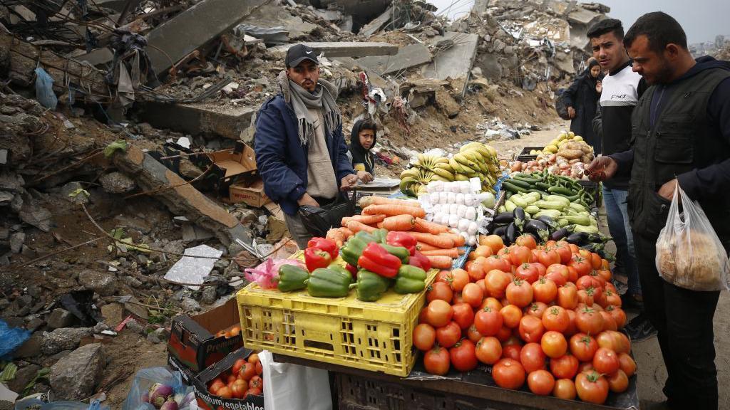 A group of Palestinians sets up a small market amid the ruins of destroyed buildings in Gaza's Jabalia refugee camp, selling fresh vegetables and fruit as daily life continues under difficult conditions following the enforcement of a ceasefire agreement on 10 February 2025.