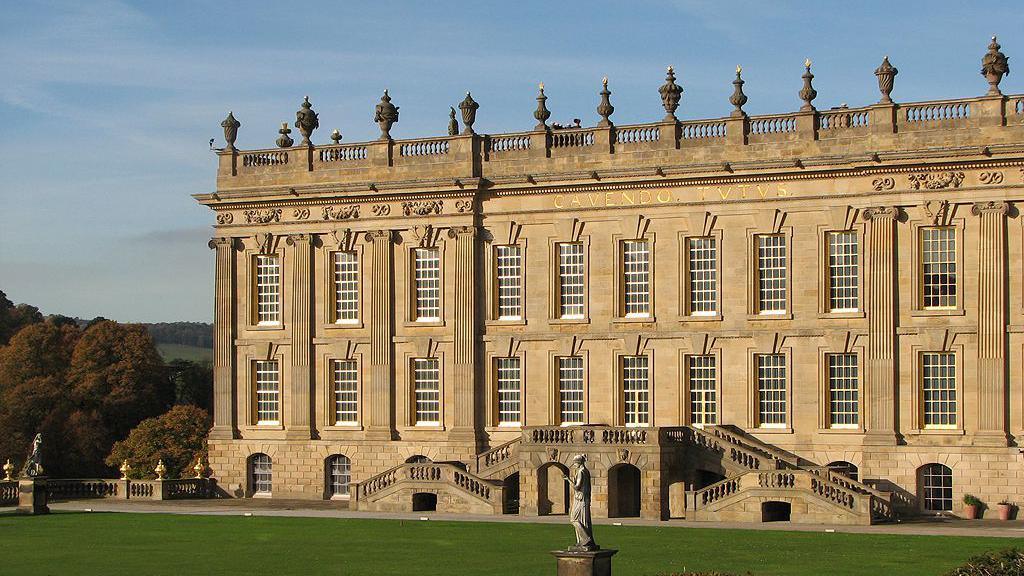 Honey-coloured classical facade of Chatsworth House in sunlight with hills in backdrop and classcial statues on lawn in front.
