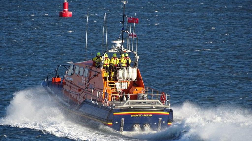 Barrow RNLI's blue and orange lifeboat travelling in the sea to the rescue. Sex people in yellow RNLU uniform and helmets are standing on the boat, which has Barrow Lifeboat written on it in yellow. 