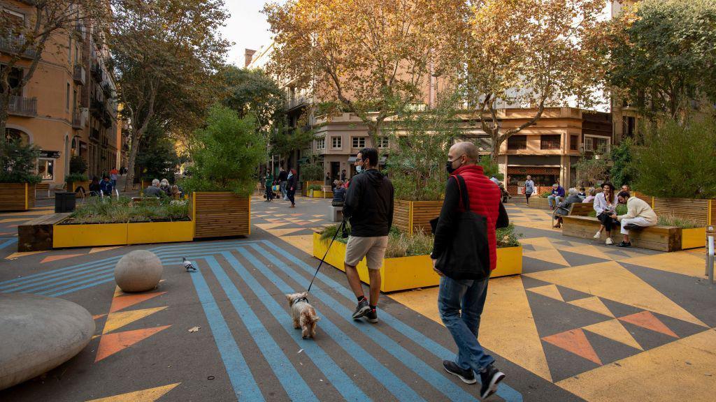 People sit on benches as others walk their dogs through an area that used to be a road but is now taken over by trees and flowerboxes. The road has been decorated in brightly coloured yellow and oranges triangles and blue stripes.