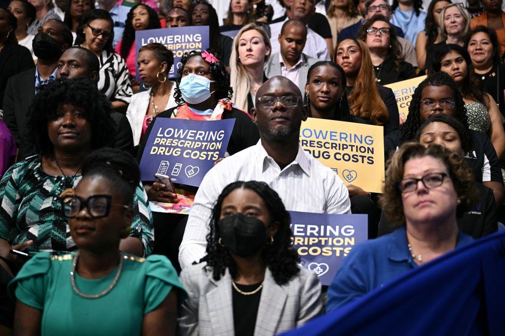 People in the crowd hold signs that read "Lowering prescription drug costs"