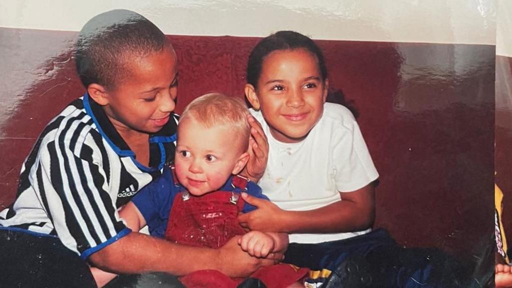 An old photograph from a disposable camera. The image shows three young children; Tyler on the left, a blonde-haired baby boy in the middle and a girl on the right. They are embracing each other while sitting on a dark red sofa. Tyler has his hand protectively on one side of the younger boy's head.