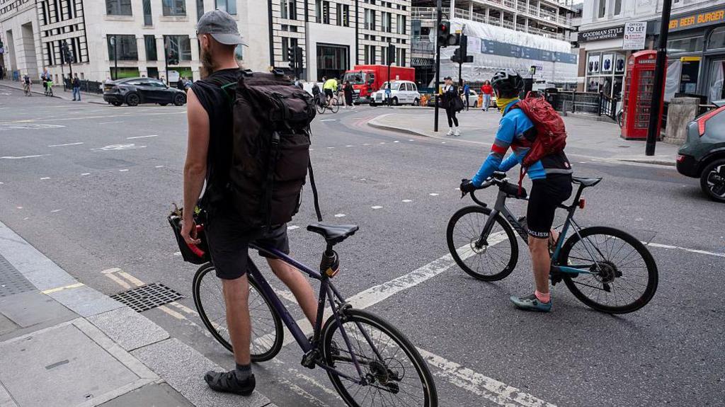 Two cyclists wait at a junction in central Londons. Cars, buses, pedestrians and a red telephone box are visible in the background.