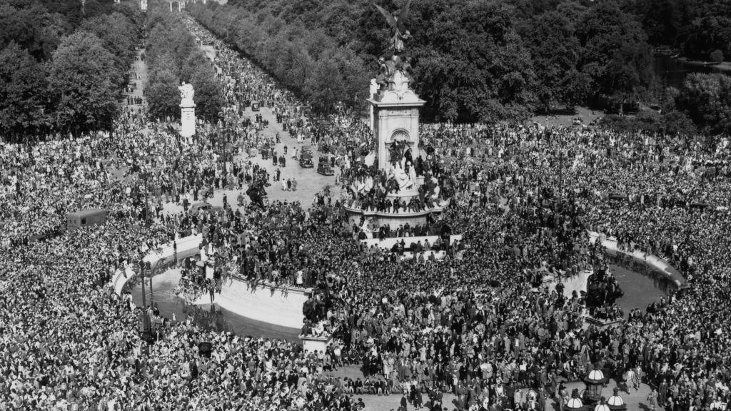 A high-angle view of crowds of people celebrating VJ Day, some climbing on the Victoria Memorial, on The Mall outside Buckingham Palace.