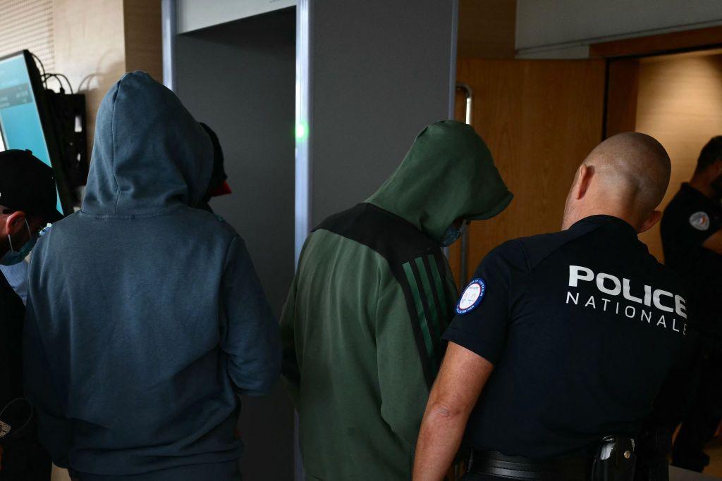 Two hooded men enter a room, flanked by a policeman