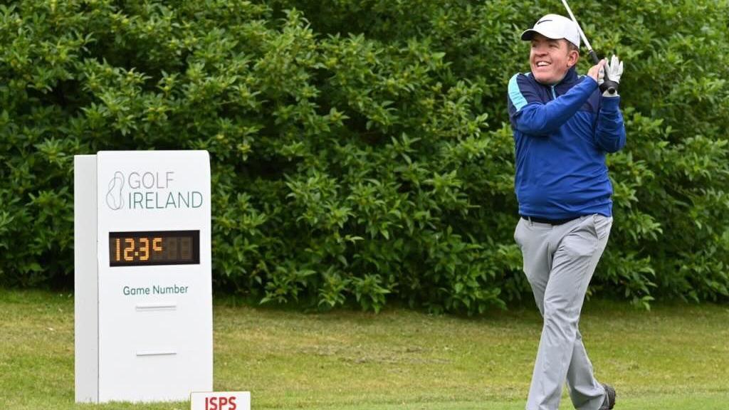 A man swinging a golf club on a golf course. He is wearing a blue cap, grey trousers and a blue long-sleeve top. He is looking into the distance as if to follow the shot. 