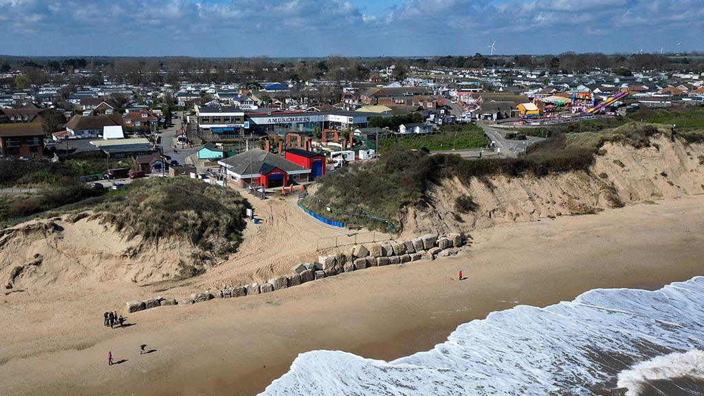 Large rocks placed outside a gap in the dunes. The lifeboat station is between the gap and the village of Hemsby can be seen in the background