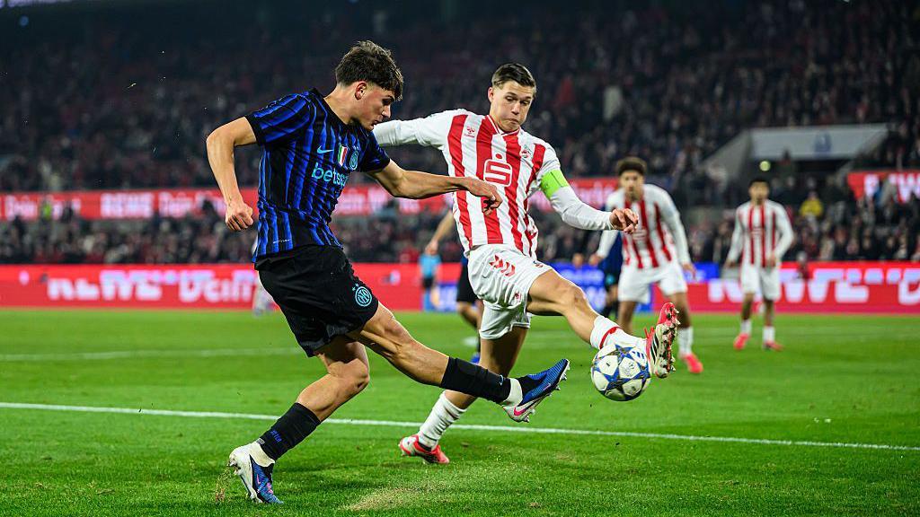 Inter Milan's Mattia Mosconi (left) kicks the ball away with his right foot as a Cologne player closes in. Inter are playing in blue-and-black striped shirts with black shorts and socks. Cologne have red-and-white striped shirts with white shorts and socks. Thousands of people can be seen in the stands behind them.