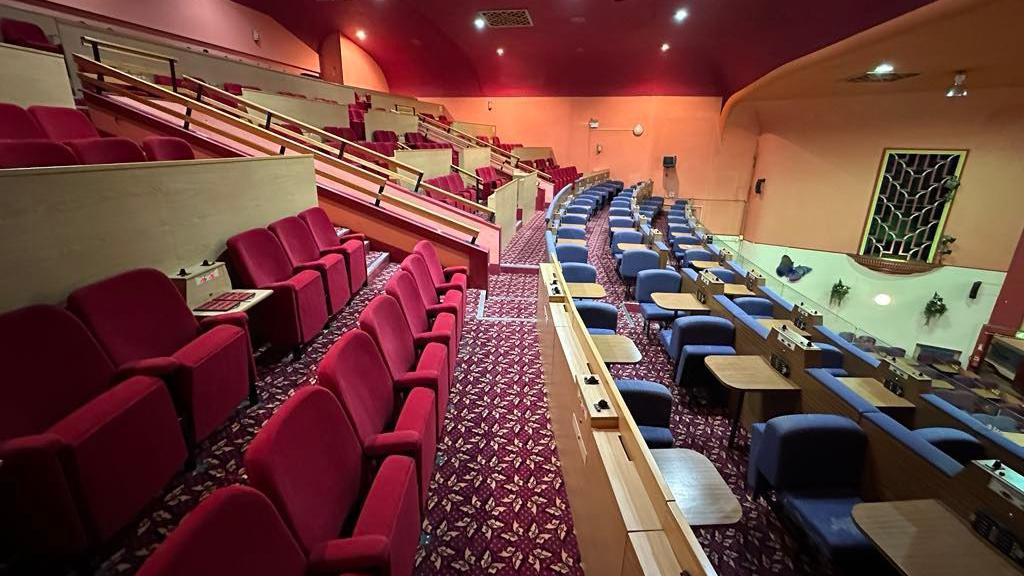 The inside of a former cinema, theatre and bingo hall, with rows of seats in a balcony area, the seats are blue and red, with a red patterned carpet, orange walls, red ceiling, and seats with some tables. 