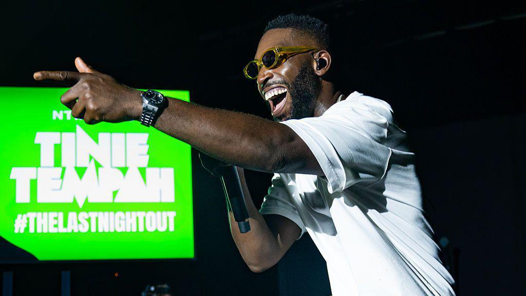 Tinie Tempah pointing left of the camera. He's smiling and wearing sunglasses and a white t-shirt while singing into a mic. Behind him is a green screen that says "Tinie Tempah - hashtag the last night out".