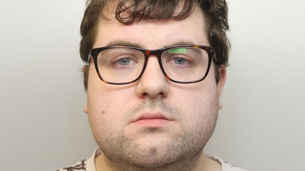 A custody mugshot of Nathan Bennett. He has short brown hair and is wearing glasses