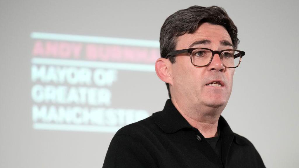 Andy Burnham stands in front of a screen with his name and title during a press conference