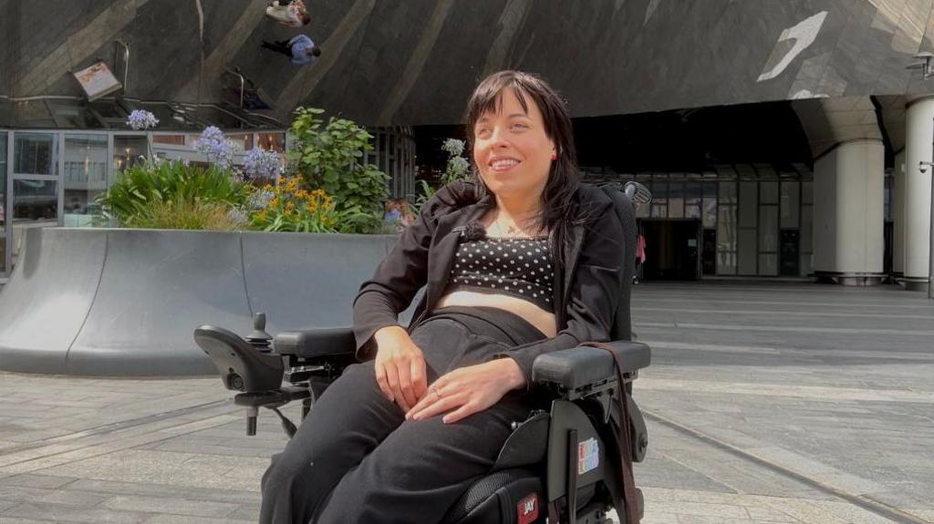 Woman sitting in a wheelchair in all black outfit smiling and looking at the camera. 