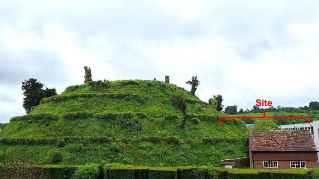 A large, green man-made mound clearly shaped like a cone, but looking like it has been done in stages due to a path cut around it. To one side is a red-brick building with pitched roof, part of it cut slightly into the bottom of the mound.