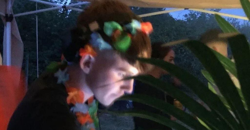 Calum Leighton pictured at a music event, with his head bent forward looking at music equipment. He is wearing a black t-shirt and a garland of flowers around his neck and in his hair. 