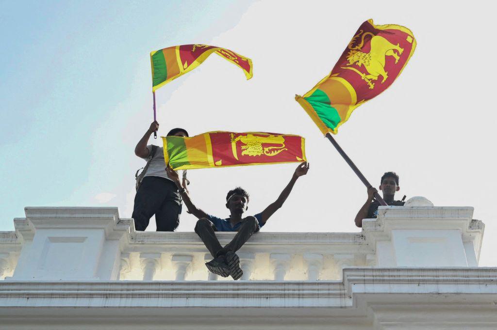 Three young men wave Sri Lankan fans from atop a white concrete balustrade