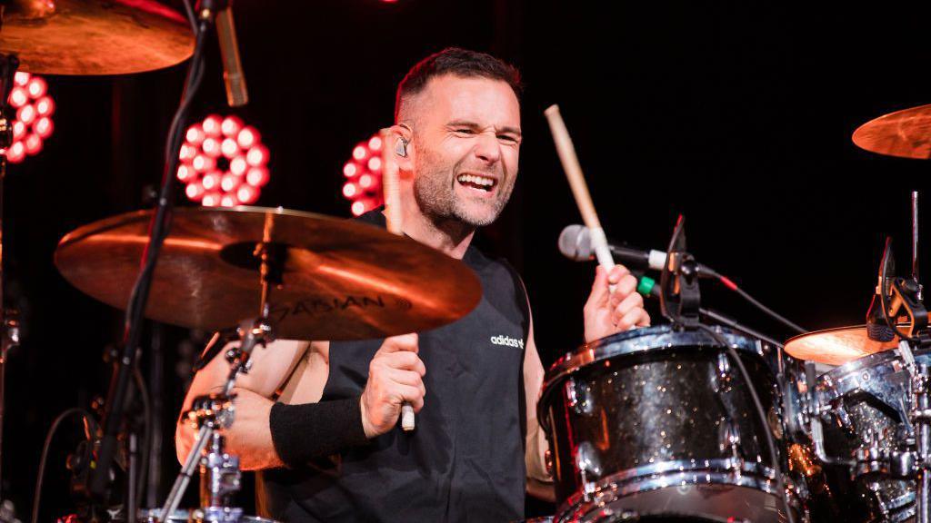 Mcfly's Harry Judd sits on stage playing the drums. He is wearing a black vest that reads "Adidas" and a black sweatband on his right wrist. He has a grimacing look on his face. He is sat in front of red stage lights.