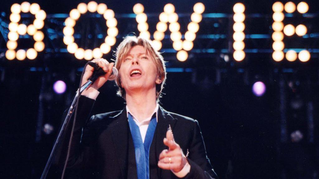 David Bowie, wearing a suit jacket and blue and white shirt, performs on stage with a sign saying BOWIE behind him 